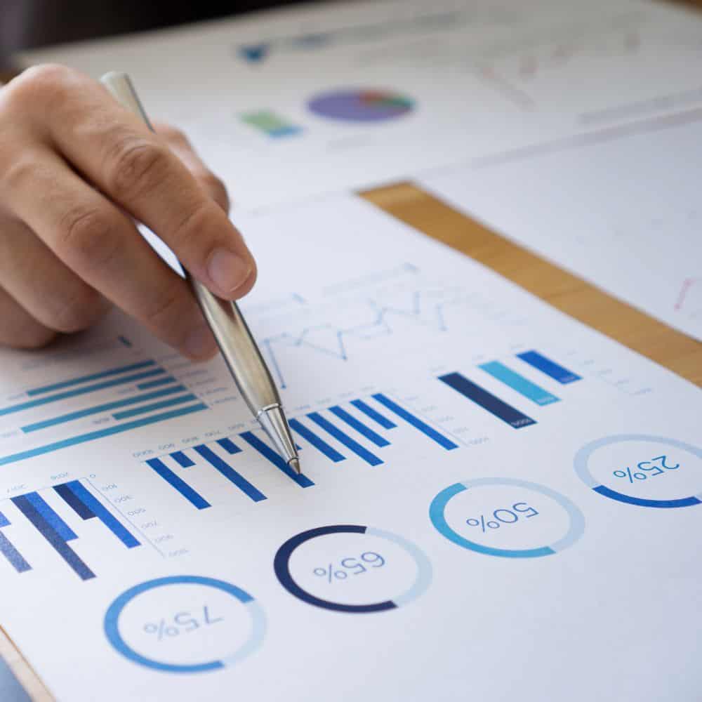 Hand holding a pen over paper with graphs and charts.