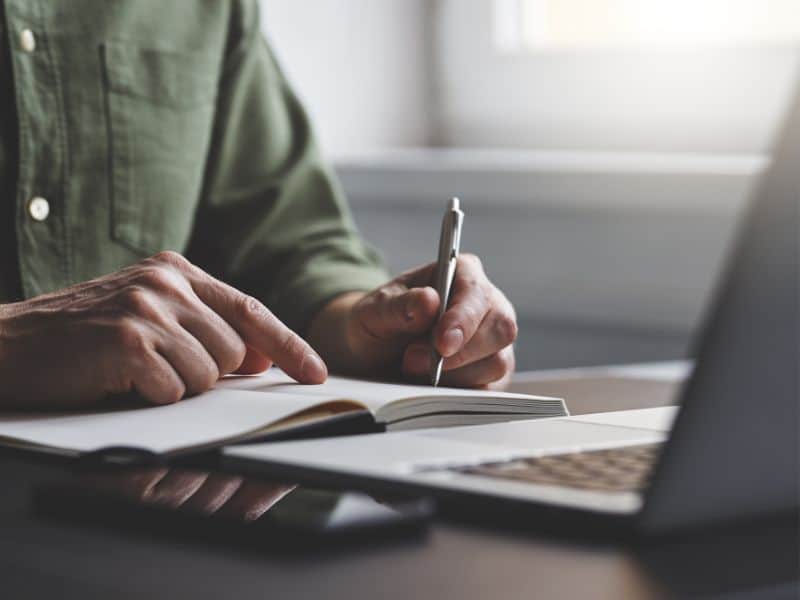 Man writing into notebook with laptop in front of him.