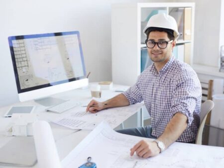 Design engineer wearing hard hat sits at a desk smiling with designs on the computer and in the foreground