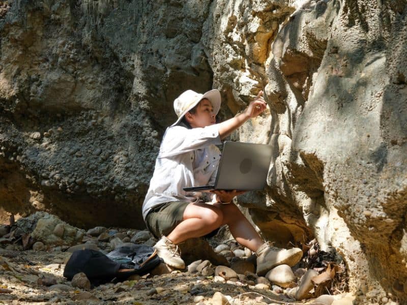 Female geologist studying a rock face and recording on a laptop