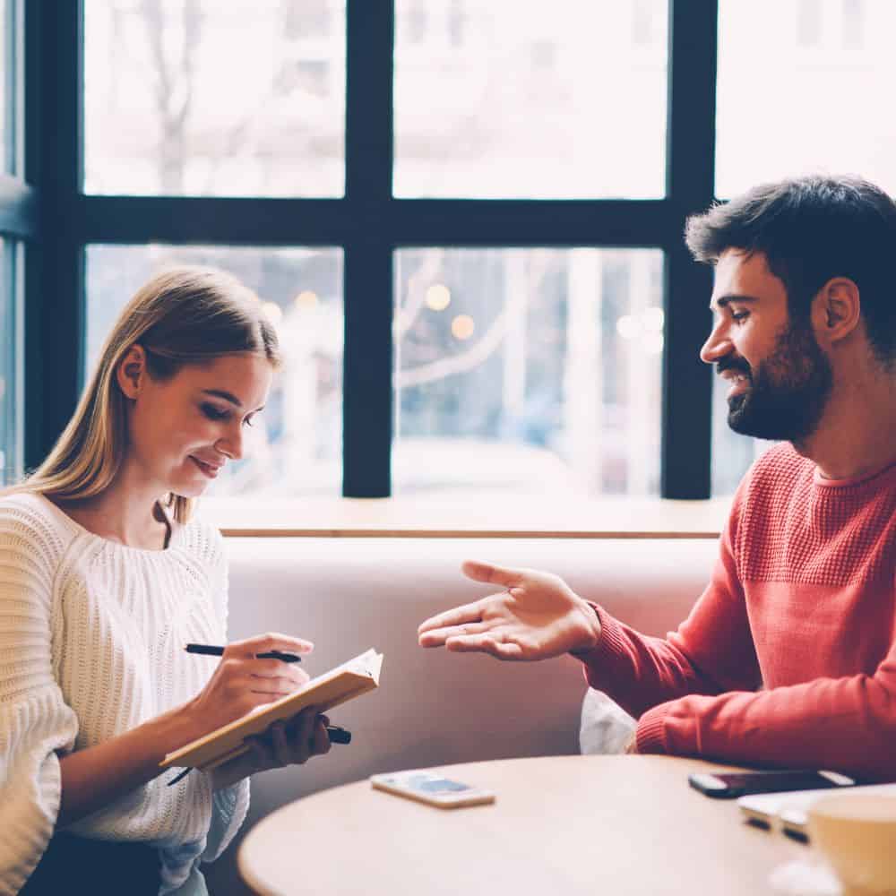 Two people at a table next to a window talking - the woman is writing notes