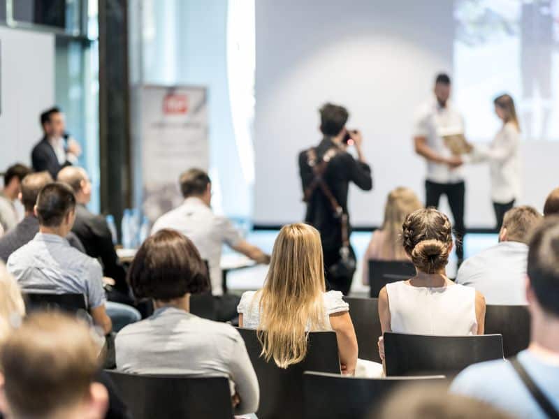 Room full of people with somebody being presented an award on stage, and being photographed