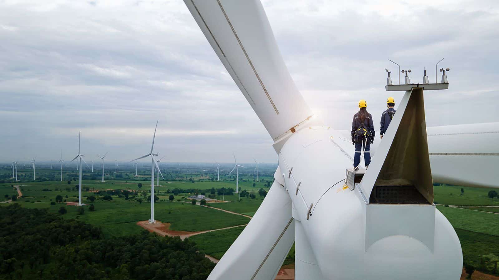 Two technicians stand on top of a wind turbine looking out onto a wind farm