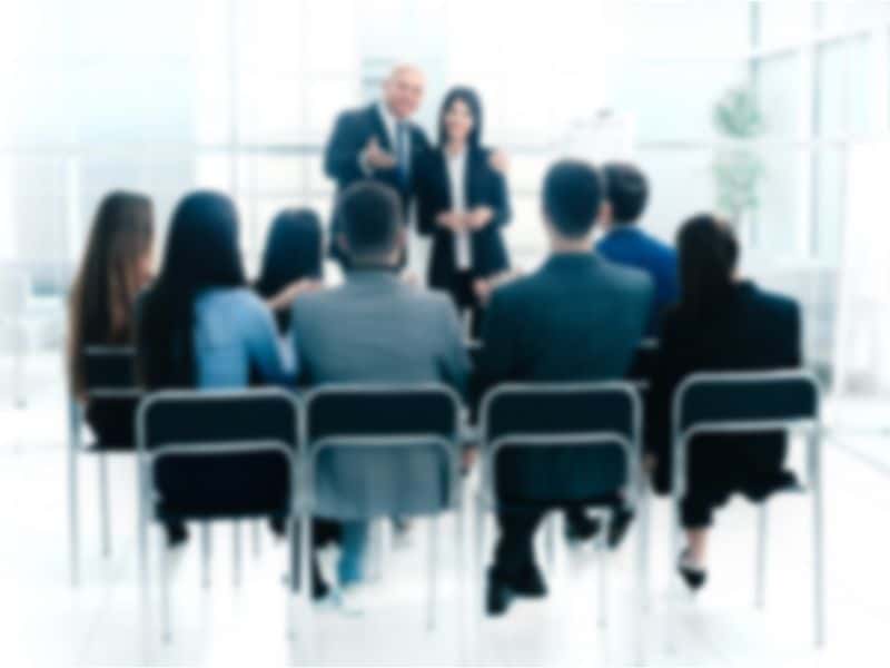 Blurred image of the backs of a group of people listening to a talk by two people standing up