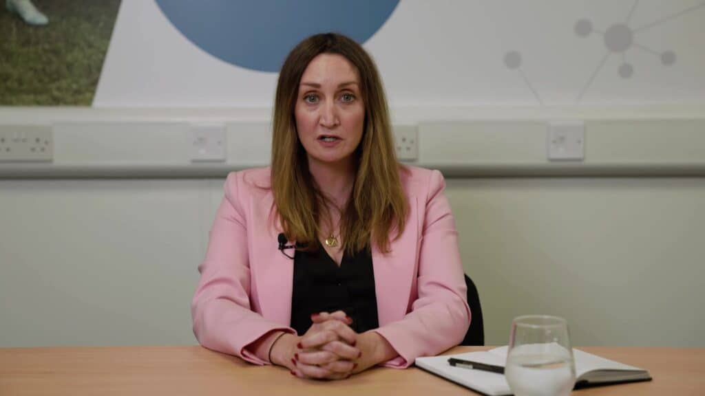 Photo of woman in a pink jacket and black shirt talking to the camera in a meeting room