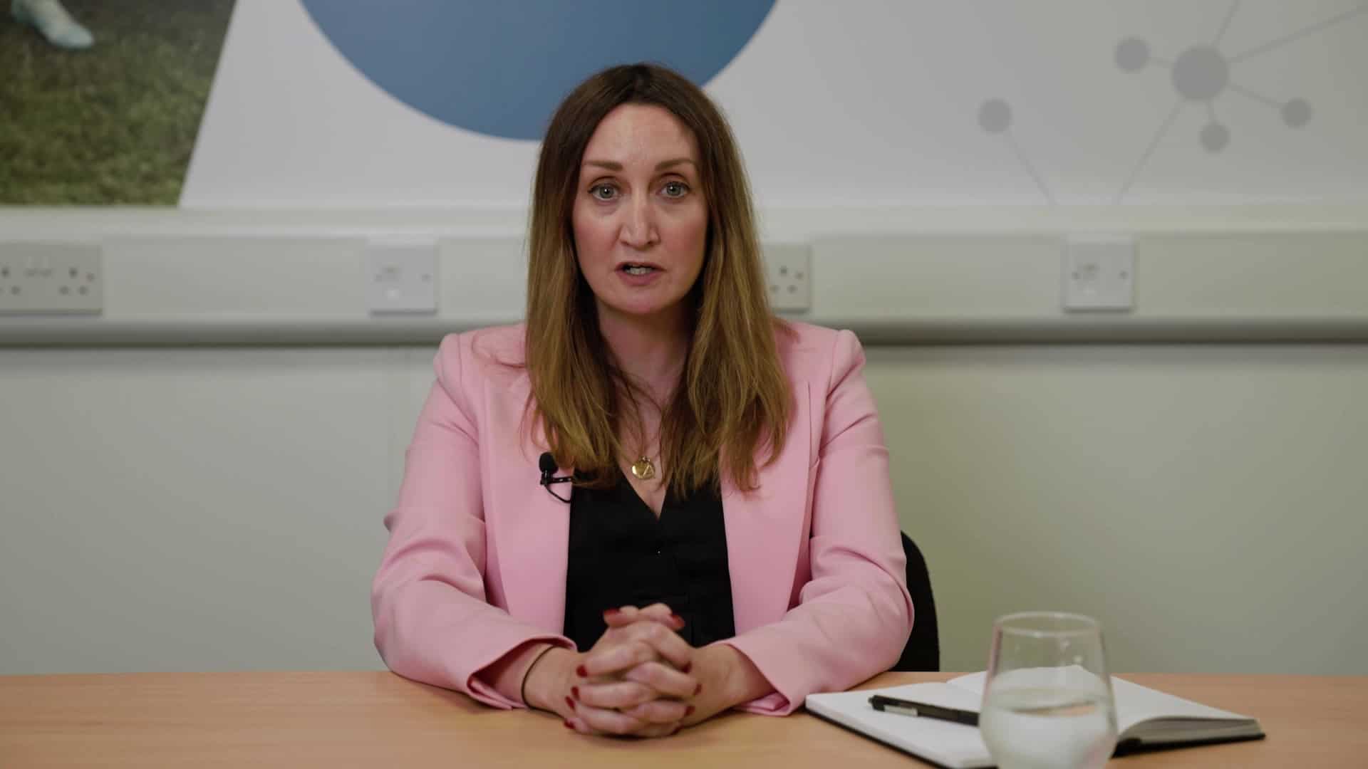 Photo of woman in a pink jacket and black shirt talking to the camera in a meeting room