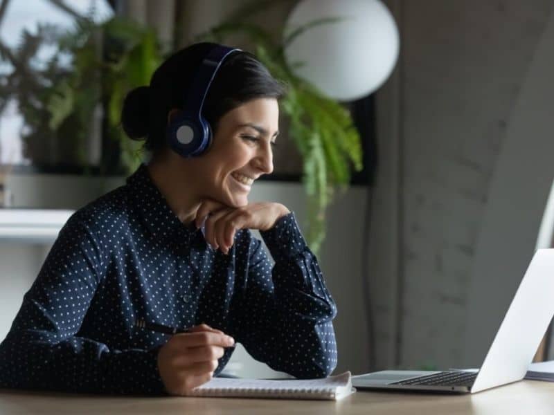 Woman wearing a headset and taking notes whilst looking at her laptop and smiling