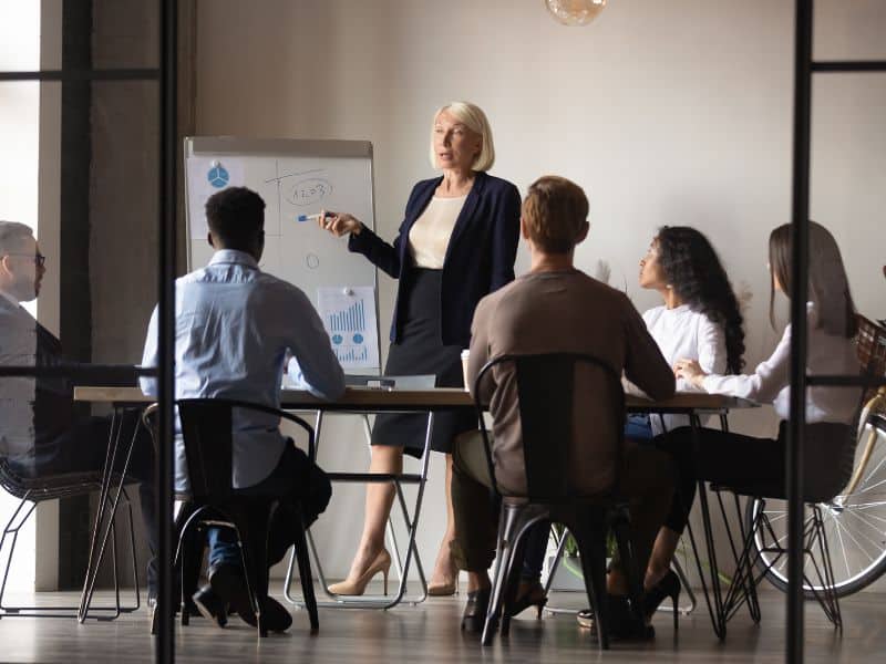 Women standing in a meeting room presenting from a white board to a group of individuals sitting down