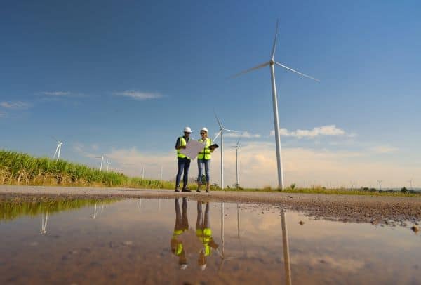 Two people standing next to a field with wind turbines in the background. They are wearing high visibility jackets and hard hats.