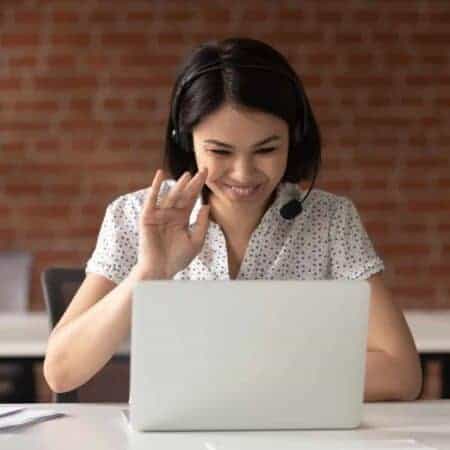 Photo of a woman smiling and waving to a computer screen, with a headset on
