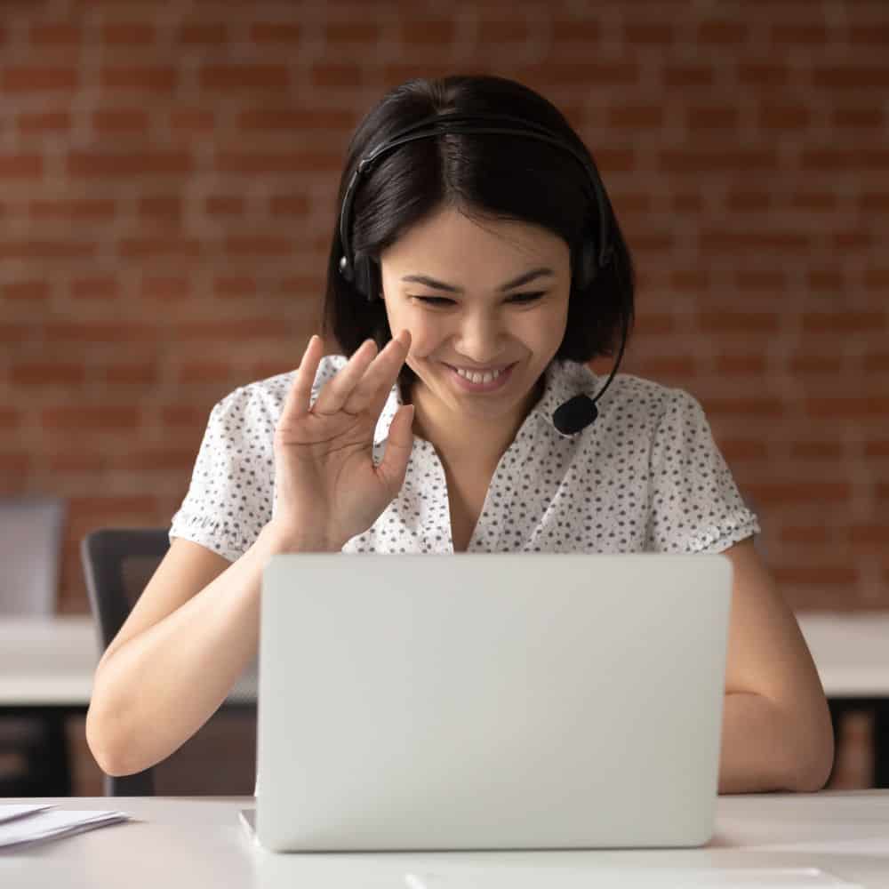 Photo of a woman smiling and waving to a computer screen, with a headset on