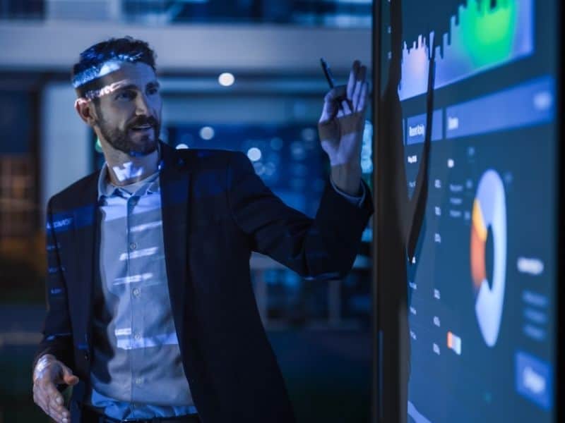 Man presenting data on a big screen. he is wearing a shirt and jacket.