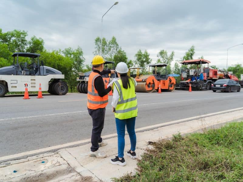 Two people standing on the side of a road looking at machinery. Both are wearing high-visibility jackets and hard hats