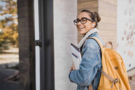 Photo of a young girl carrying a laptop and notebooks and walking into a building. she has a yellow rucksack on her back