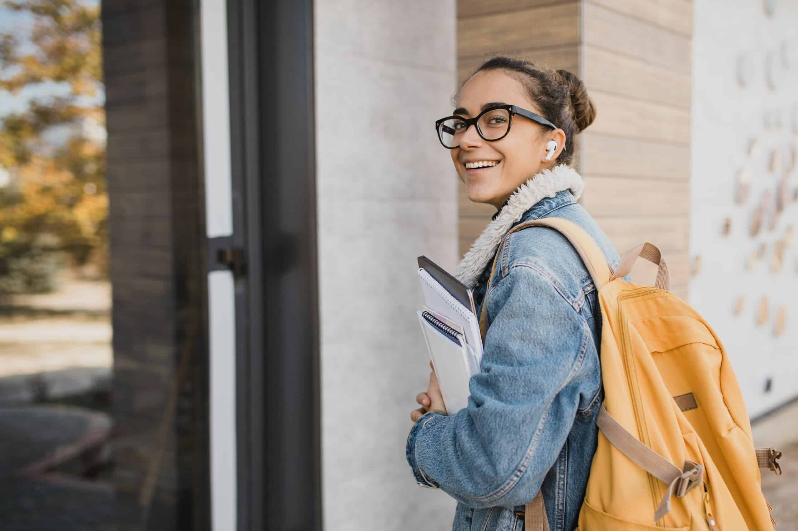 Photo of a young girl carrying a laptop and notebooks and walking into a building. she has a yellow rucksack on her back