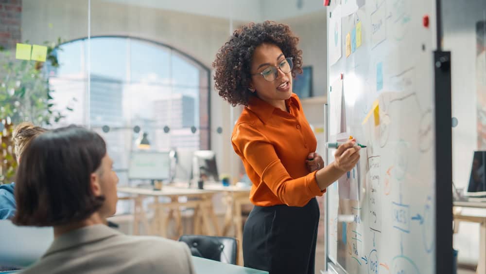 Woman standing at a board and writing on it, other people are watching