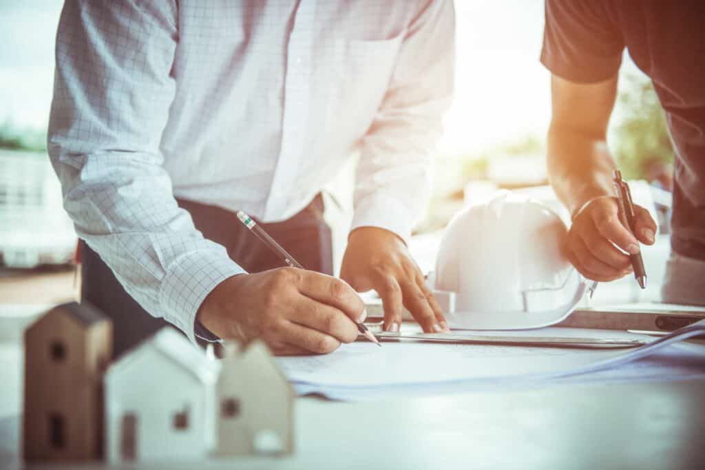 The torsos of two people standing over a desk annotating a sheet. there is a hard hat and model houses on the table