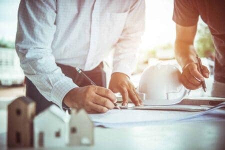 The torsos of two people standing over a desk annotating a sheet. there is a hard hat and model houses on the table