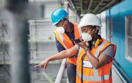 Two people in high-visibility jackets and hard hats. One is pointing at something in a warehouse/factory.
