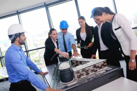 Group of people looking at a building model on a table. Some people are wearing hard hats
