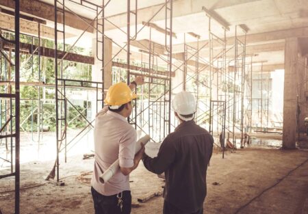 Two people in a building site, wearing hard hats and looking at the construction work