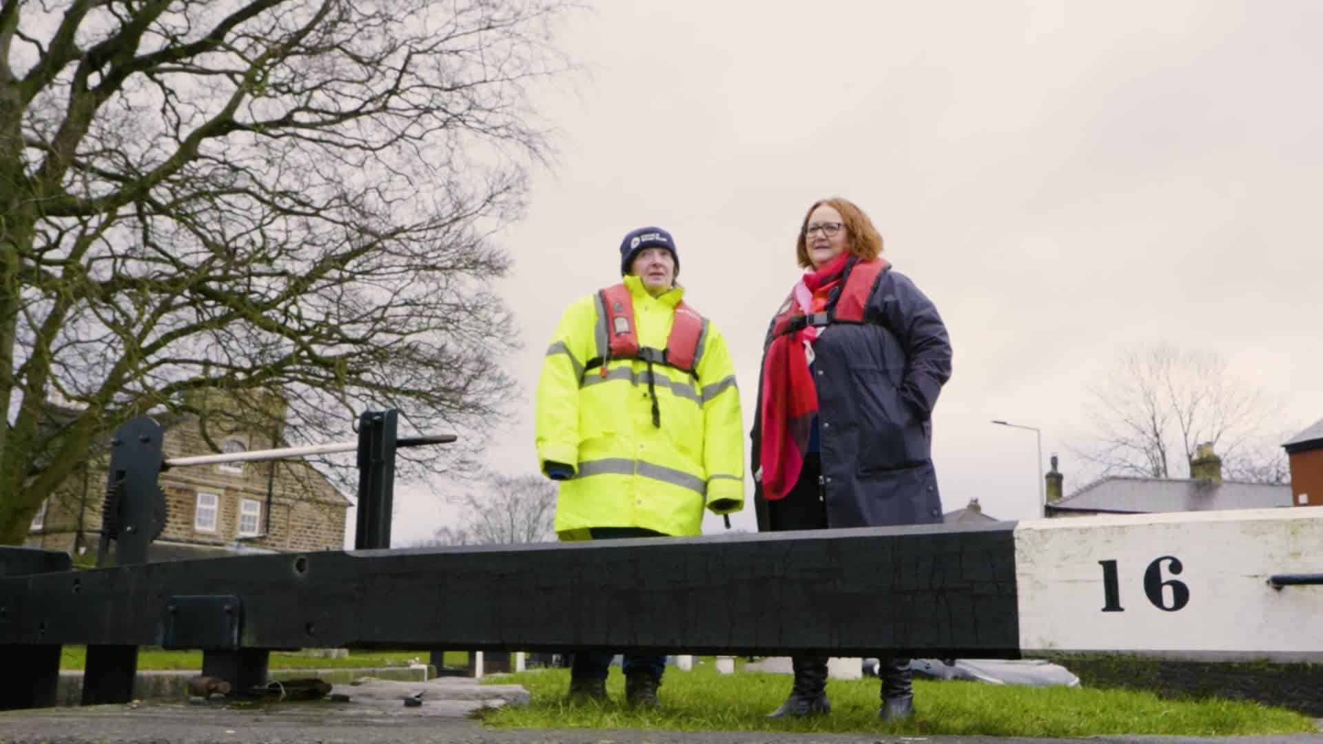 Two people stood by a lock in the UK. one is wearing a high-visibility coat