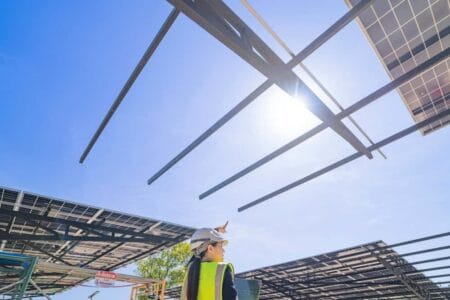 A woman underneath a solar panel structure and pointing up at the sky to the sun