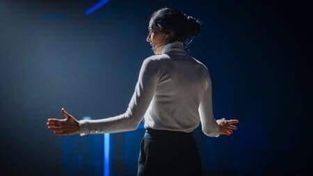 Woman with her back to the shot, with arms open wide and speaking to an audience