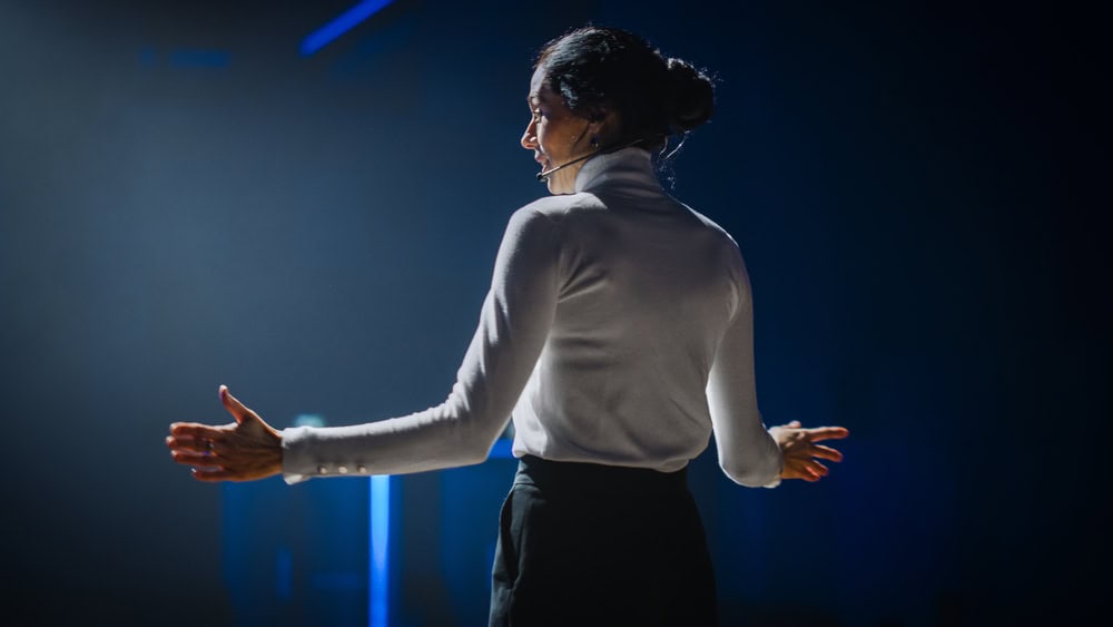 Woman with her back to the shot, with arms open wide and speaking to an audience