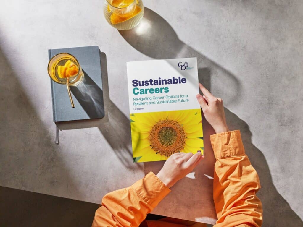 A picture of a desk with a notepad, a drink of water and a person's arms holding a Liz Painter's book, titled: 'Sustainable Careers: Navigating Career Options for a Resilient and Sustainable Future'
