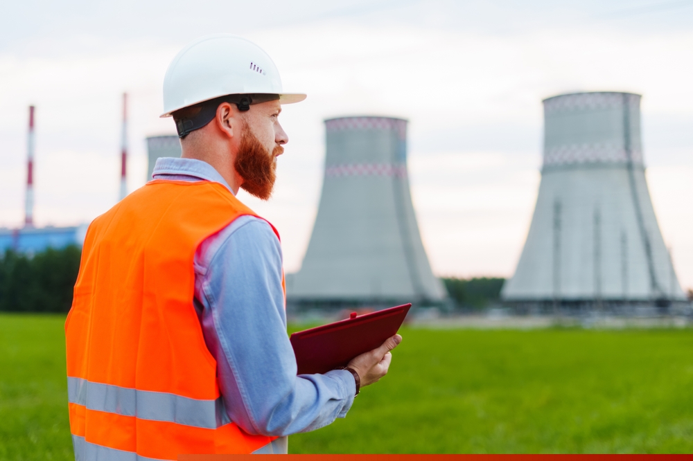 A picture of a man wearing a high vis and a hard hat, looking over at a nuclear power station and taking notes