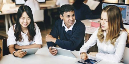 Three school students sat in a classroom, discussing a topic and looking at their tablets