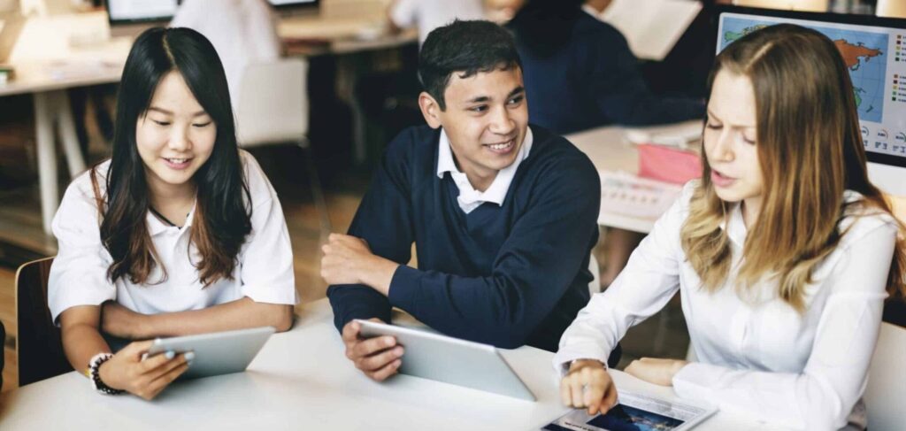 Three school students sat in a classroom, discussing a topic and looking at their tablets