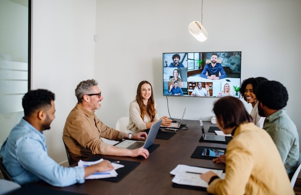 A meeting room with 6 people around a table with a screen dialled into a virtual meeting