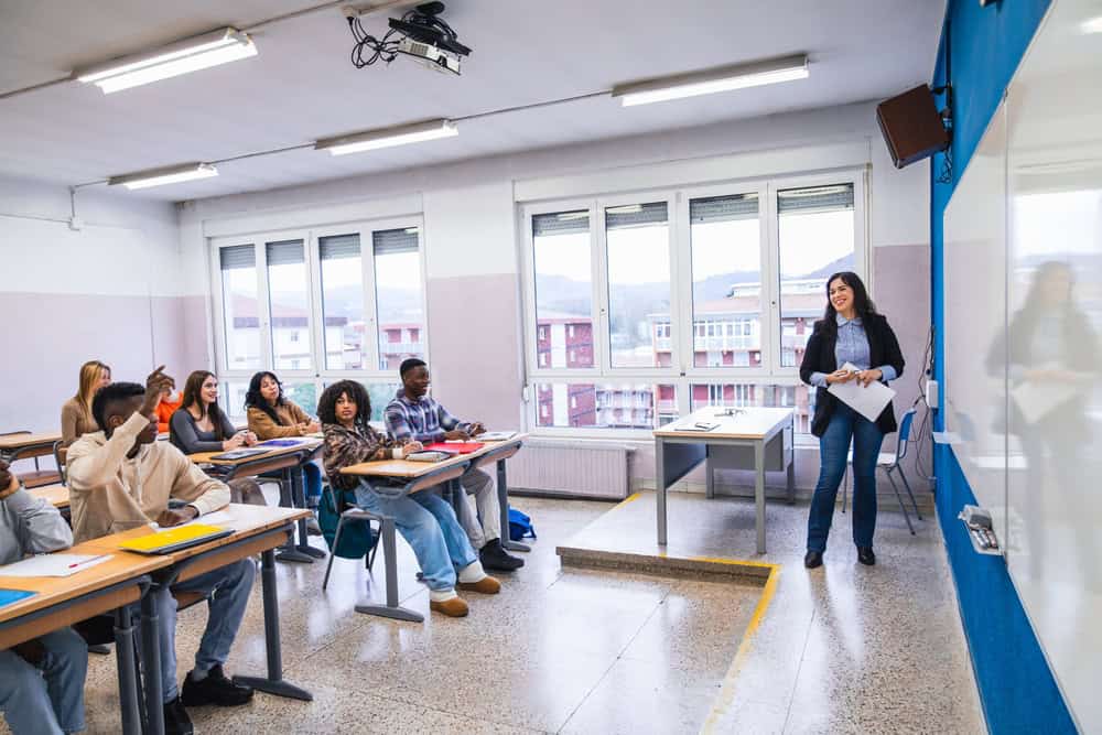 Picture of a classroom with a teacher and students where one student has their hand up to answer a question